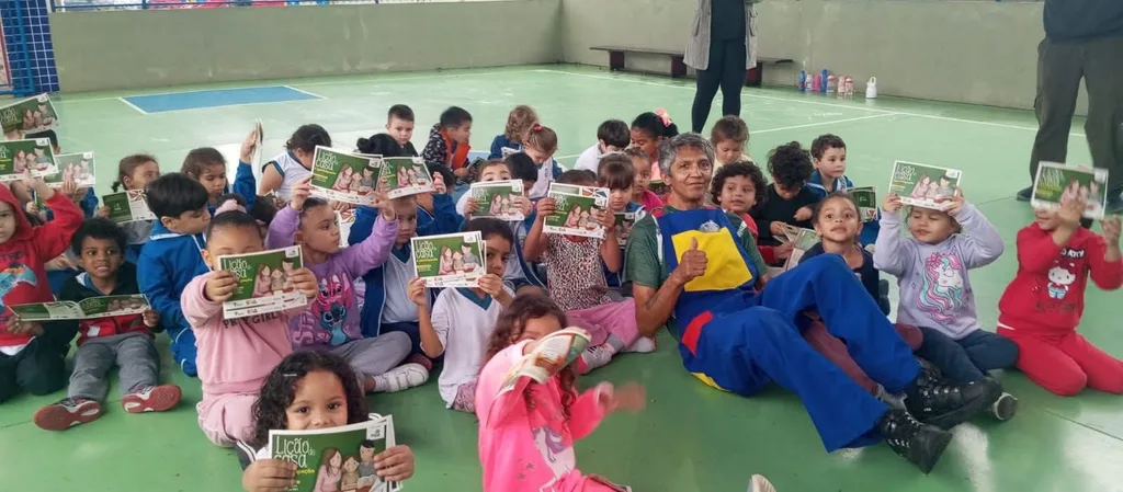 Beto sentado no chão de uma creche, cercado de crianças que seguram livros