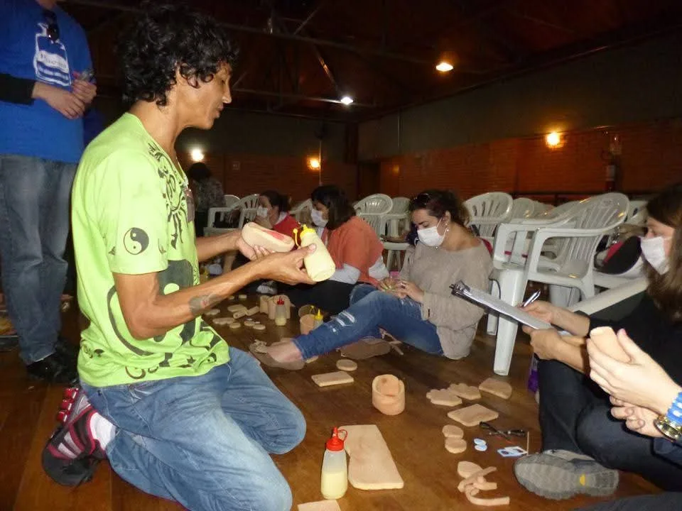 Beto ajoelhado mostrando a peça de espuma de um fantoche a educadores sentados no chão